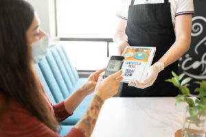Young woman scanning QR code of contactless menu through smartphone in restaurant